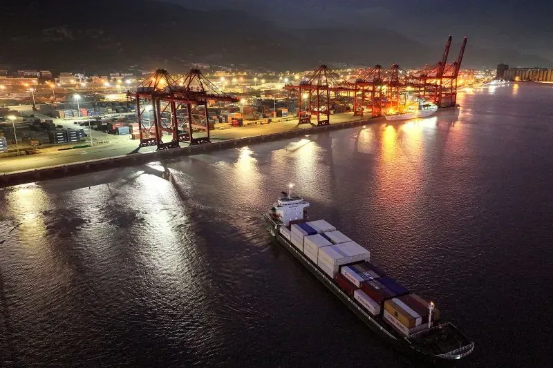 صورة لميناء بحري ليلاً، حيث تظهر سفينة شحن تحمل حاويات، مع وجود رافعات ومخازن مضاءة في الخلفية. تعكس الصورة تأثير الرسوم الجمركية على التجارة.