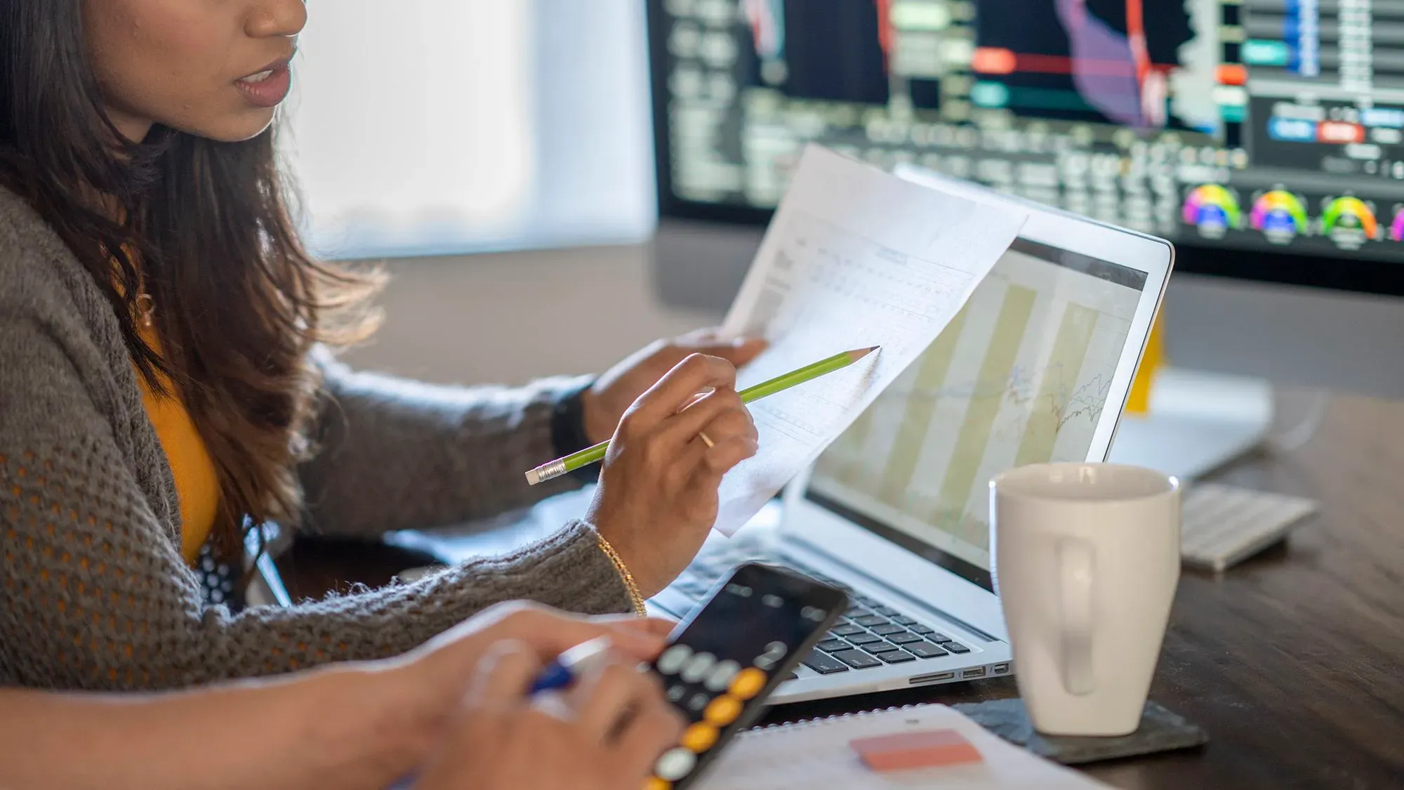 امرأة تستخدم حاسوبًا محمولًا أثناء مراجعة مستندات مالية، مع شاشة تعرض بيانات سوقية في الخلفية، تعكس أهمية الاستثمار في الأسهم الخاصة.