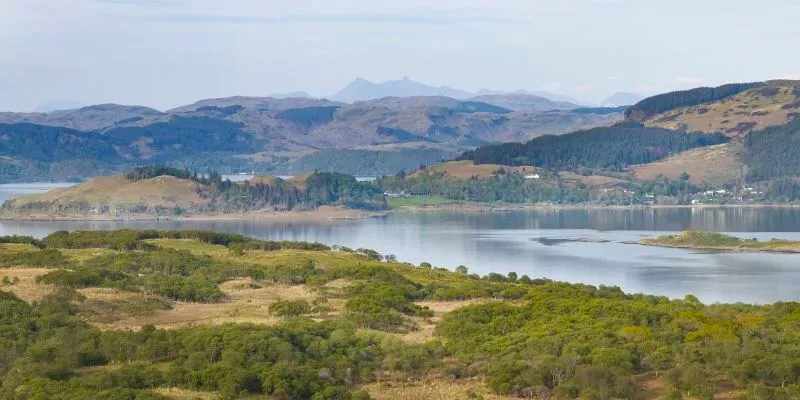 منظر طبيعي لجزيرة شونا الاسكتلندية، مع غابات ومراعي، تُظهر جمال المناظر الطبيعية المحيطة والمياه الزرقاء.