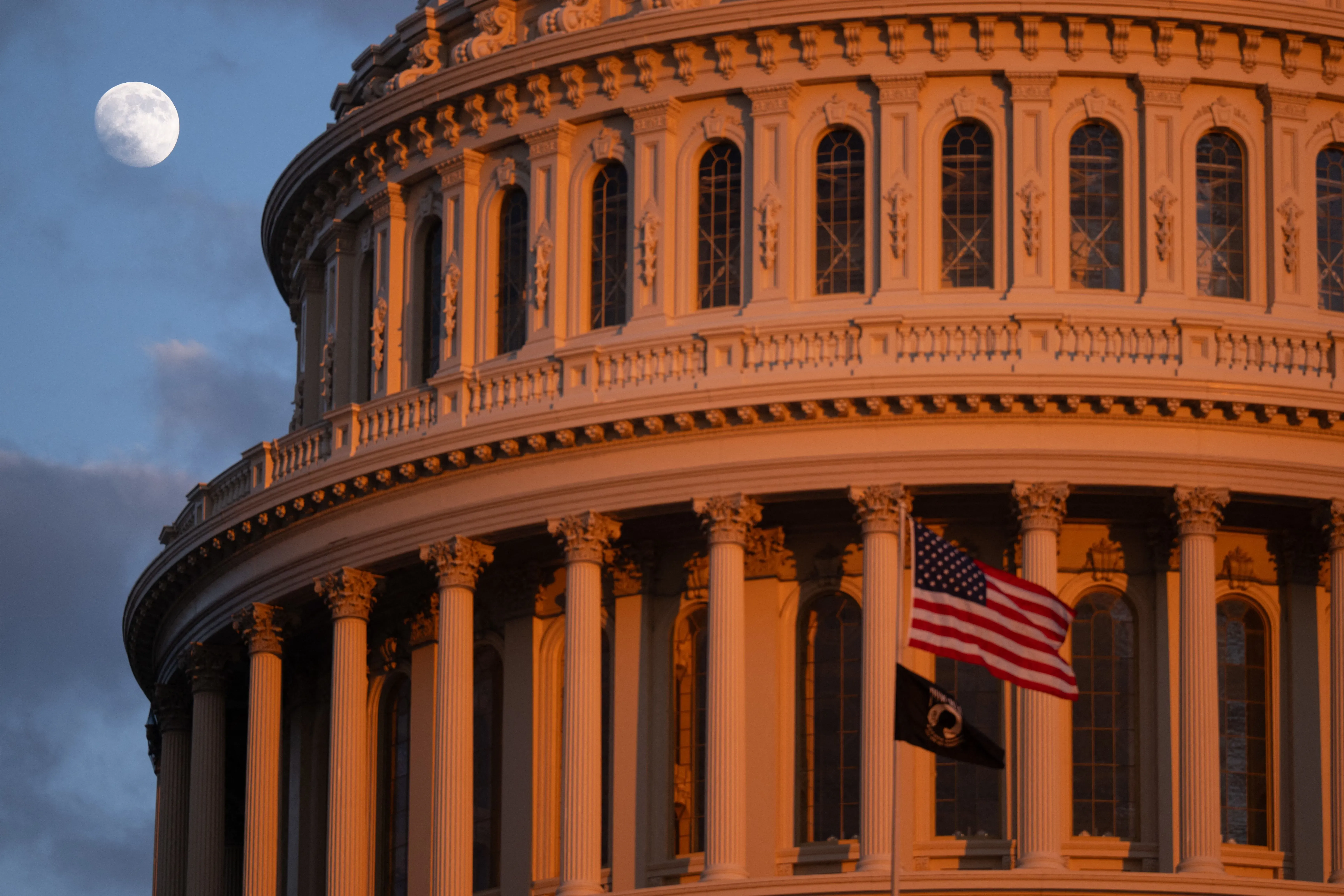 قبة مبنى الكابيتول الأمريكي تضيء بأشعة الشمس الغاربة، مع علم الولايات المتحدة يرفرف، والقمر في السماء، مما يرمز إلى أهمية السياسة في الانتخابات القادمة.