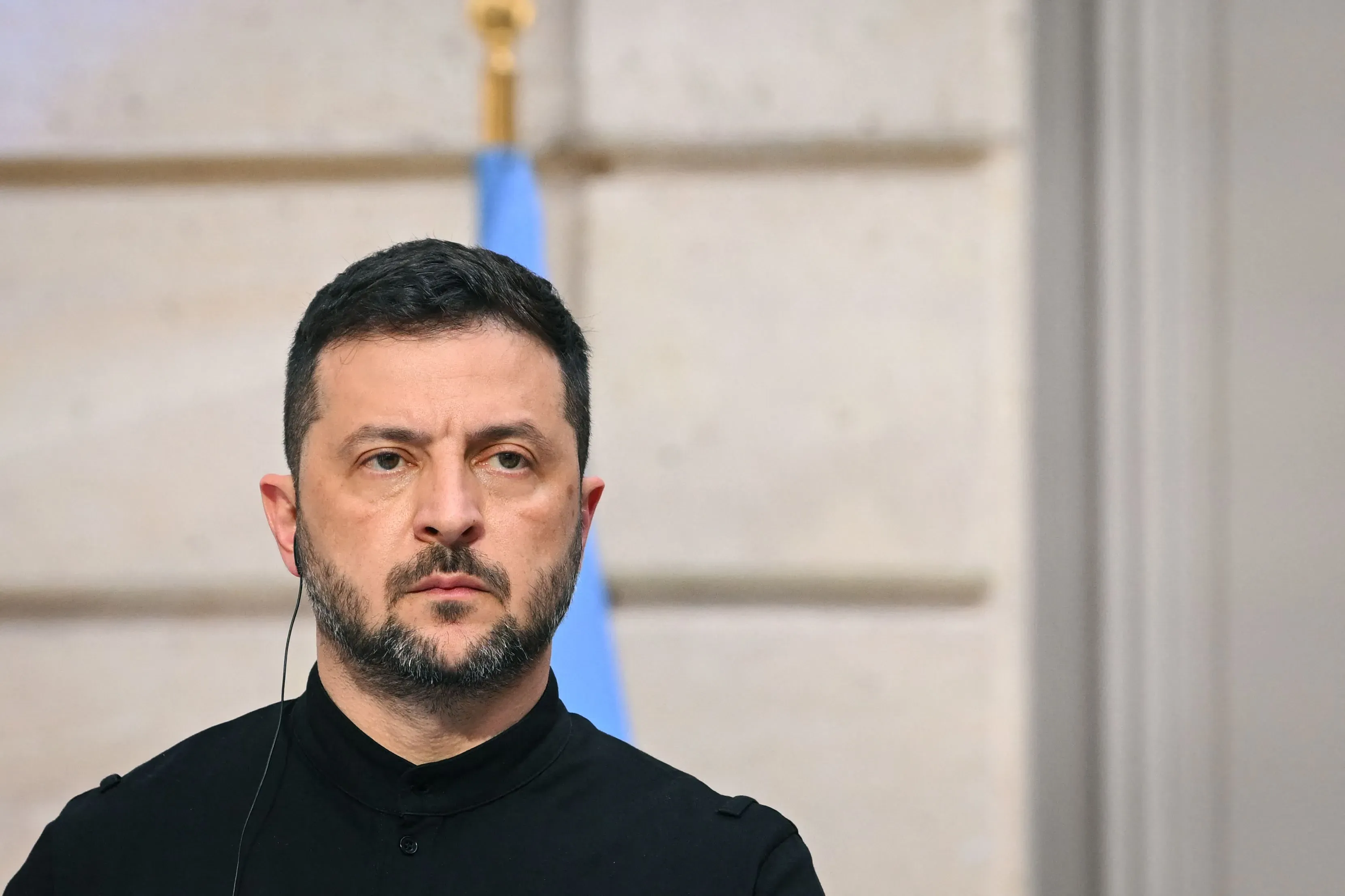 زيلينسكي، الرئيس الأوكراني، يظهر بوجه جاد خلال مؤتمر صحفي، مع العلم الأوكراني في الخلفية، مع التركيز على التحديات الأمنية التي تواجه بلاده.