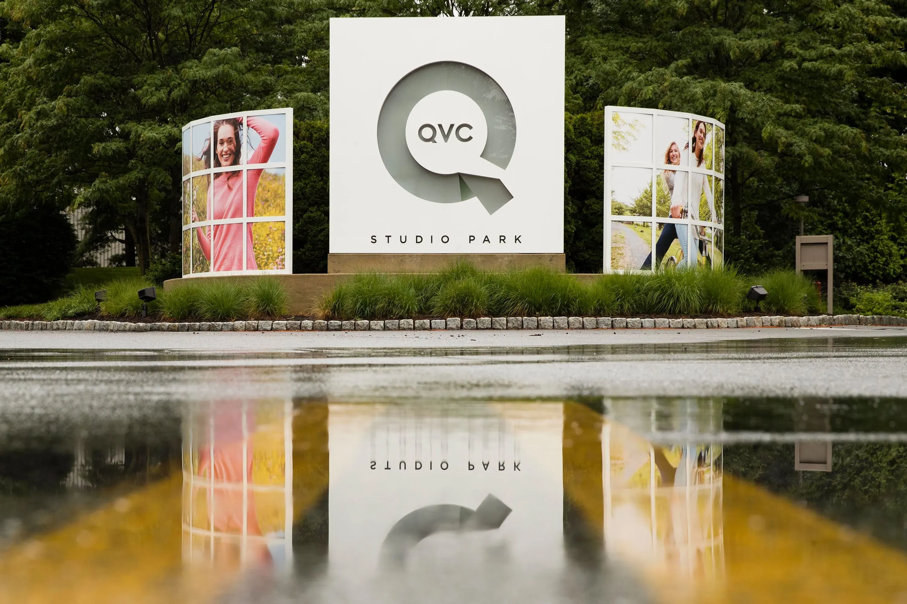 علامة QVC في حديقة الاستوديو، محاطة بصور ترويجية لموظفين، تعكس تاريخ الشركة في التسوق المباشر وتحدياتها الحالية.