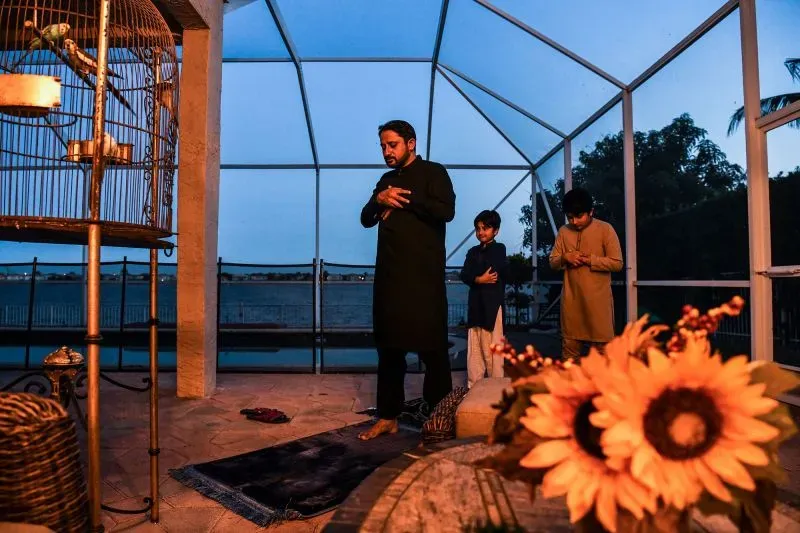 رجل وطفلان يؤدون الصلاة في وقت الغروب، مع ظهور هلال رمضان في الأفق، بينما تتزين الطاولة بأزهار دوار الشمس.