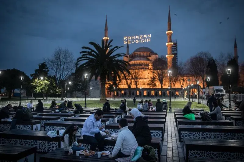 أشخاص يتناولون الطعام في حديقة قرب مسجد مضاء، مع لافتة مضيئة تروج لشهر رمضان، في أجواء رمضانية مميزة.