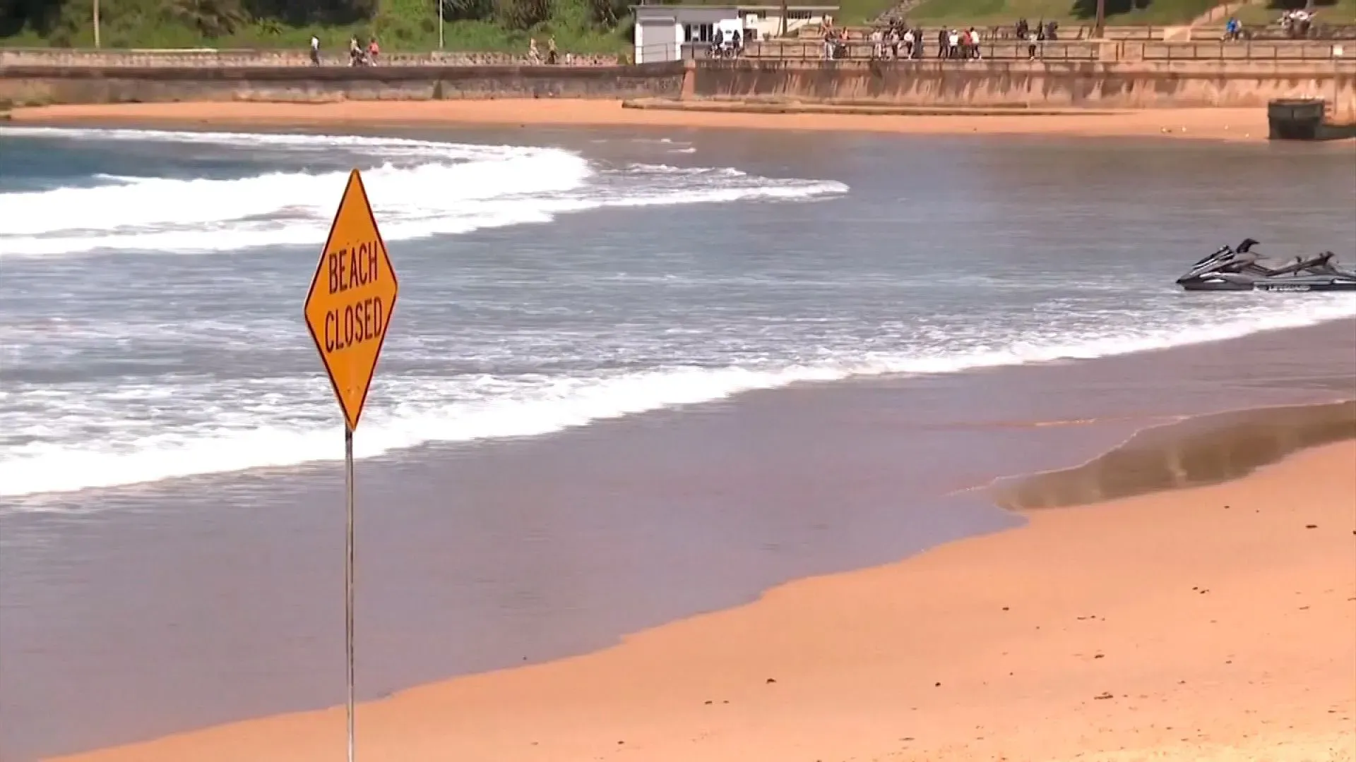 شاطئ مغلق في سيدني بعد هجوم سمكة قرش، مع لافتة تحذر الزوار من دخول المياه، وأمواج تتلاطم على الرمال.