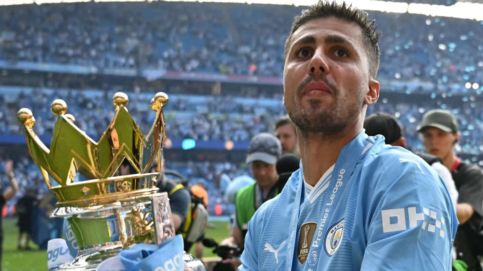 أيوب بوعدّي لاعب وسط مانشستر سيتي الشاب يحمل ميدالية الدوري الإنجليزي وكأس البطولة، مع جماهير في الخلفية.