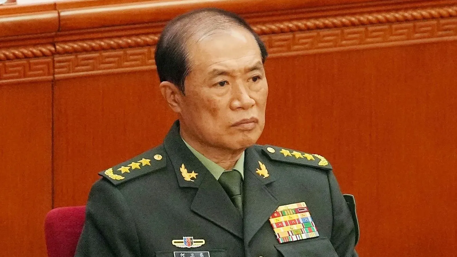 جنرال يرتدي الزي العسكري الصيني، يجلس في قاعة رسمية مع تعبير جاد، وسط خلفية خشبية، في سياق غياب ملحوظ عن حدث غرس الأشجار السنوي.