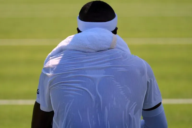 لاعب تنس يجلس على الملعب، مرتديًا قميصًا مبللاً بالعرق، مع منشفة على كتفيه، في يوم حار خلال بطولة ويمبلدون.