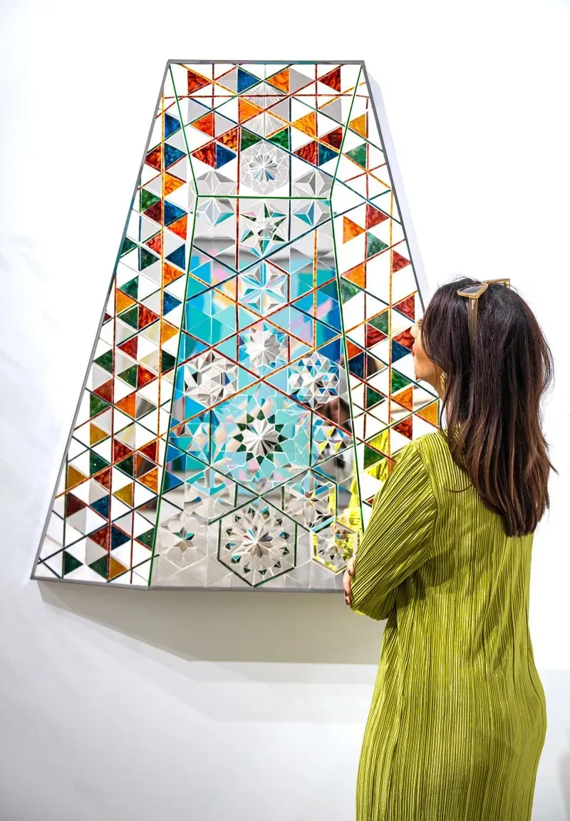 امرأة ترتدي ملابس خضراء تتأمل عملًا فنيًا معقدًا من الزجاج الملون والمرايا، يعكس تفاصيل فنية غنية تعبر عن الثقافة العربية.
