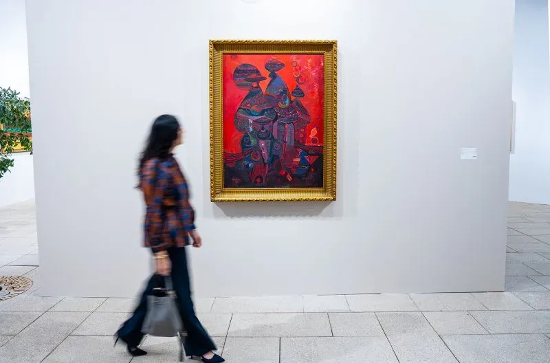 امرأة تسير بجانب لوحة فنية ملونة في إطار ذهبي، تعرض ألوانًا دافئة ونقوشًا معقدة، في معرض فني بالمملكة العربية السعودية.