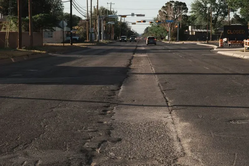 شارع متضرر في منطقة سكنية، تظهر فيه حفر وتشققات على الأسفلت، مما يعكس الحاجة إلى تحسينات في البنية التحتية والنقل.