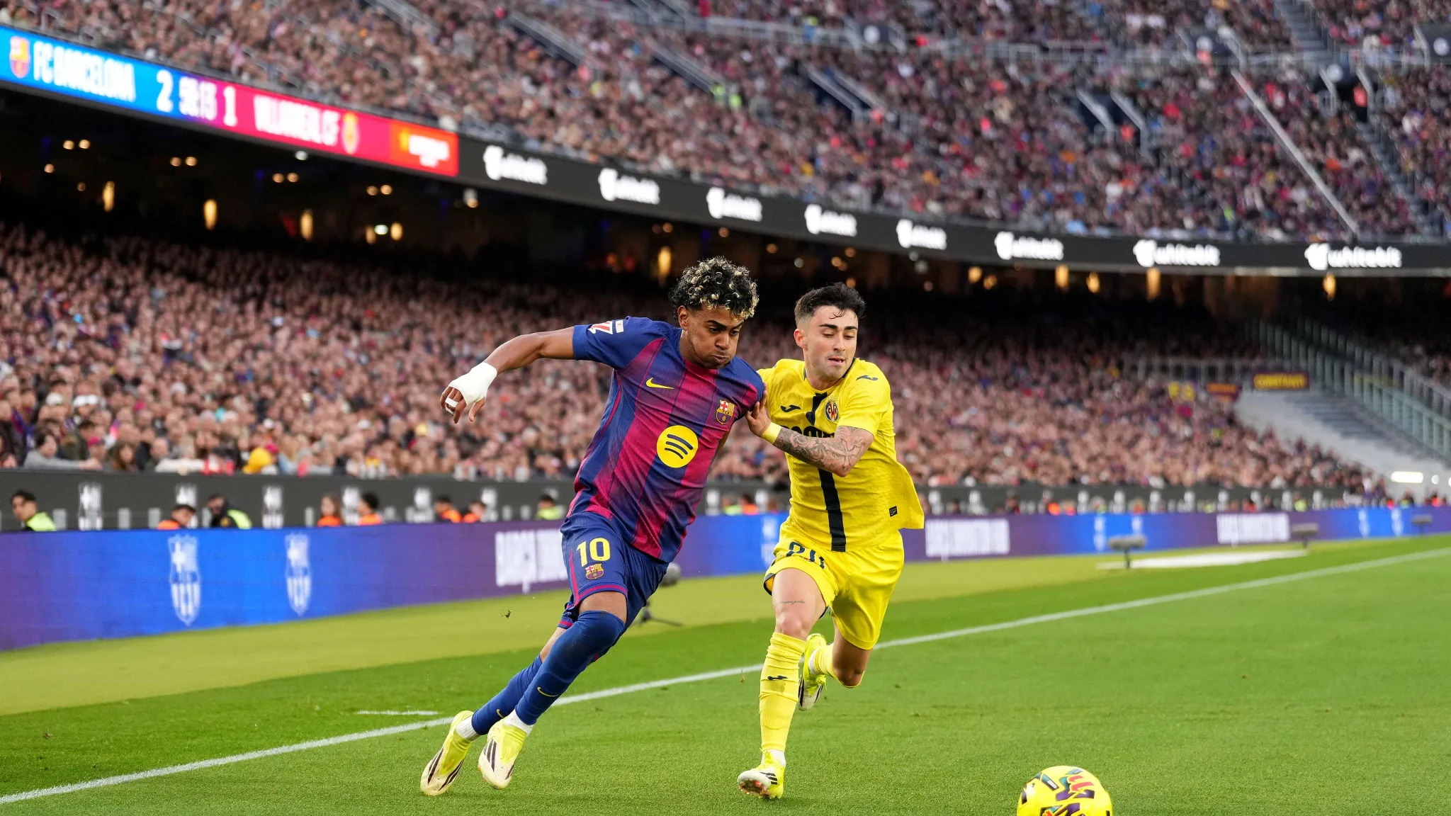 لاعب برشلونة لامين يامال يتقدم بالكرة أمام مدافع في مباراة حماسية أمام جمهور كبير في ملعب كامب نو.