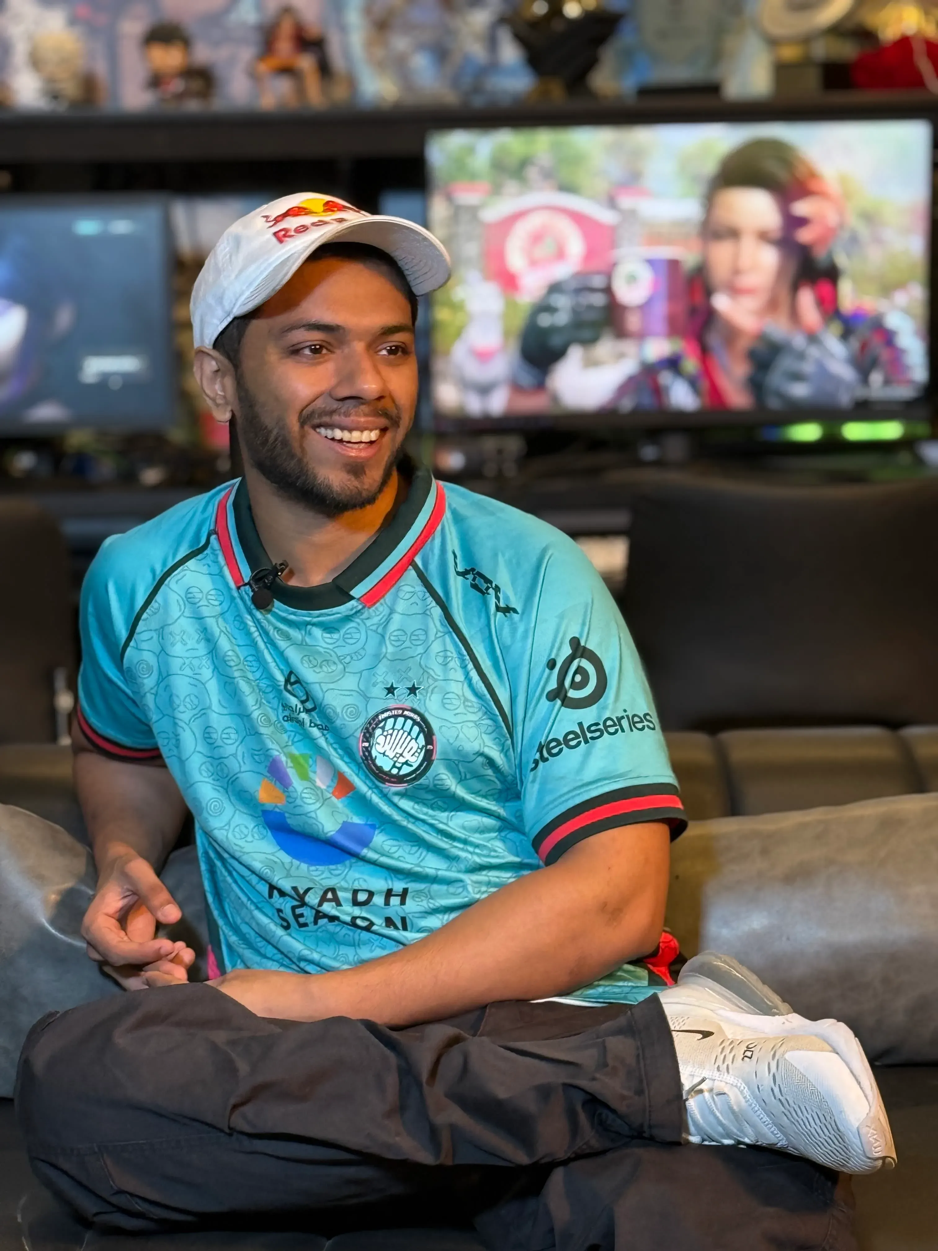 أرسلان "آش" صديق، بطل Tekken الباكستاني، مبتسم في صالة ألعاب، يرتدي قميص فريقه، محاطًا بجوائزه ووحدات التحكم.