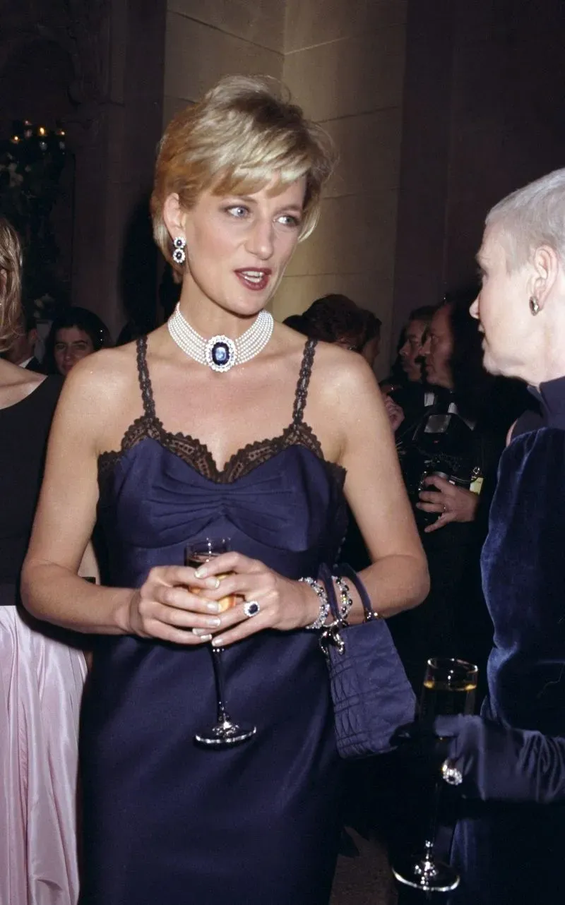 ديانا، أميرة ويلز، تتحدث مع سيدة أخرى في حفل الميت غالا عام 1996، مرتدية فستانًا داكنًا مزينًا باللؤلؤ والياقوت.