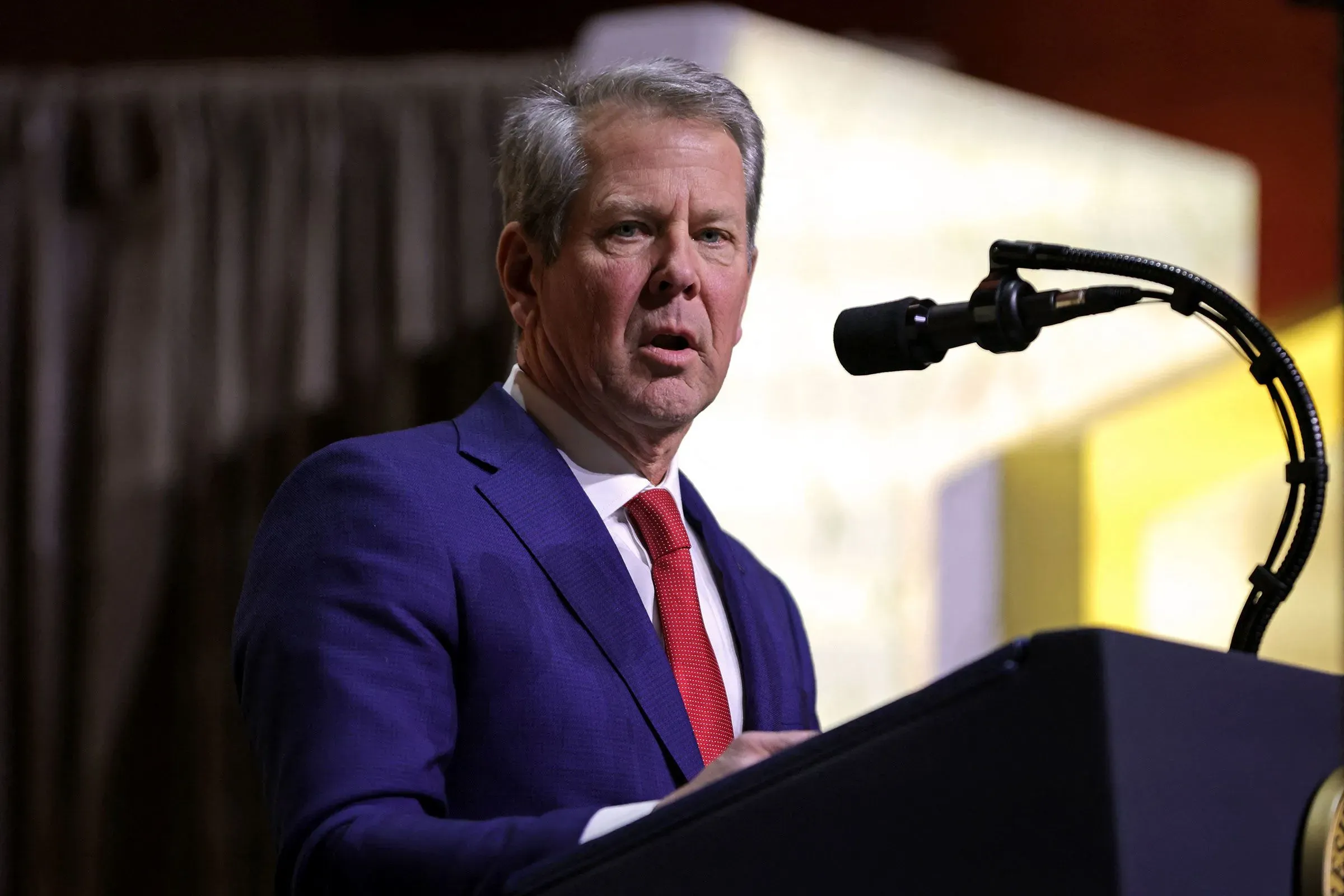 رجل يتحدث من منصة، مرتديًا بدلة زرقاء وربطة عنق حمراء، خلال حدث سياسي، مع خلفية غير واضحة.