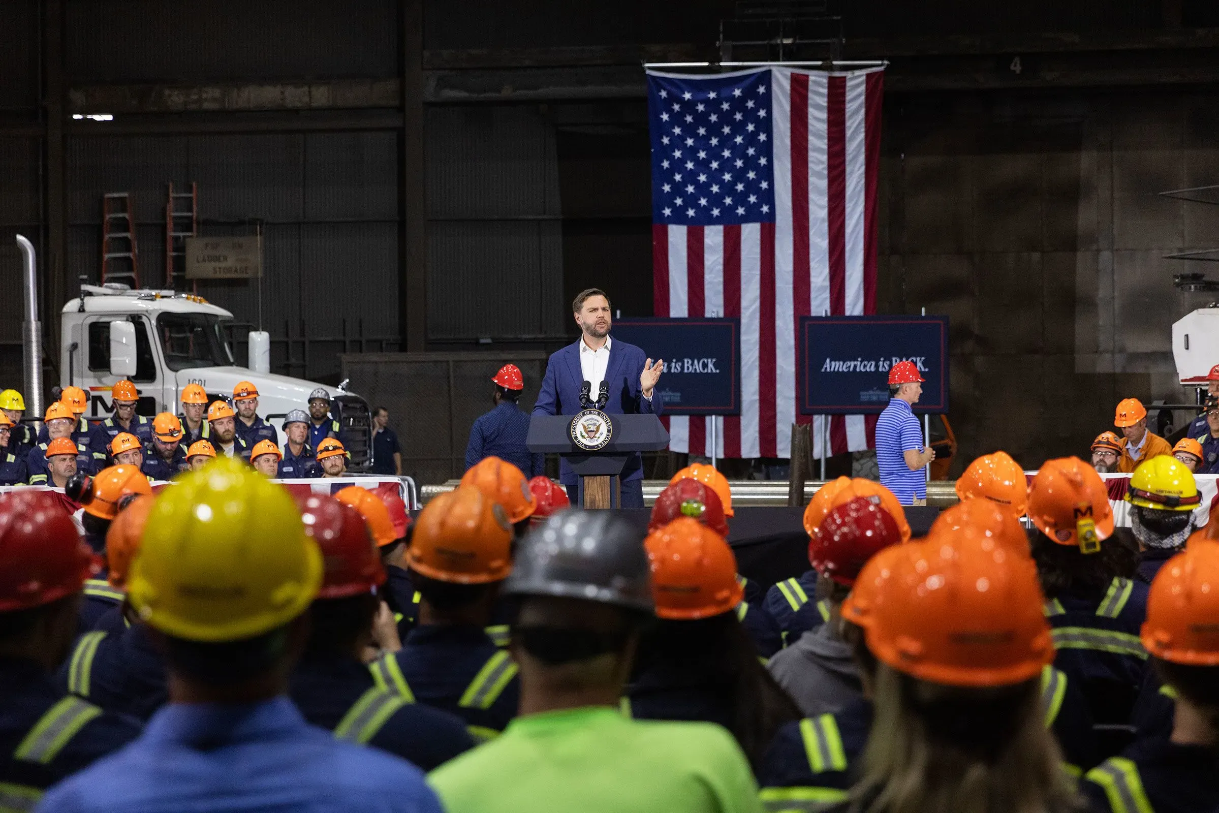 خطاب لجمهوري يتحدث أمام حشد من العمال في موقع صناعي، مع وجود لافتات وأعلام أمريكية خلفه.