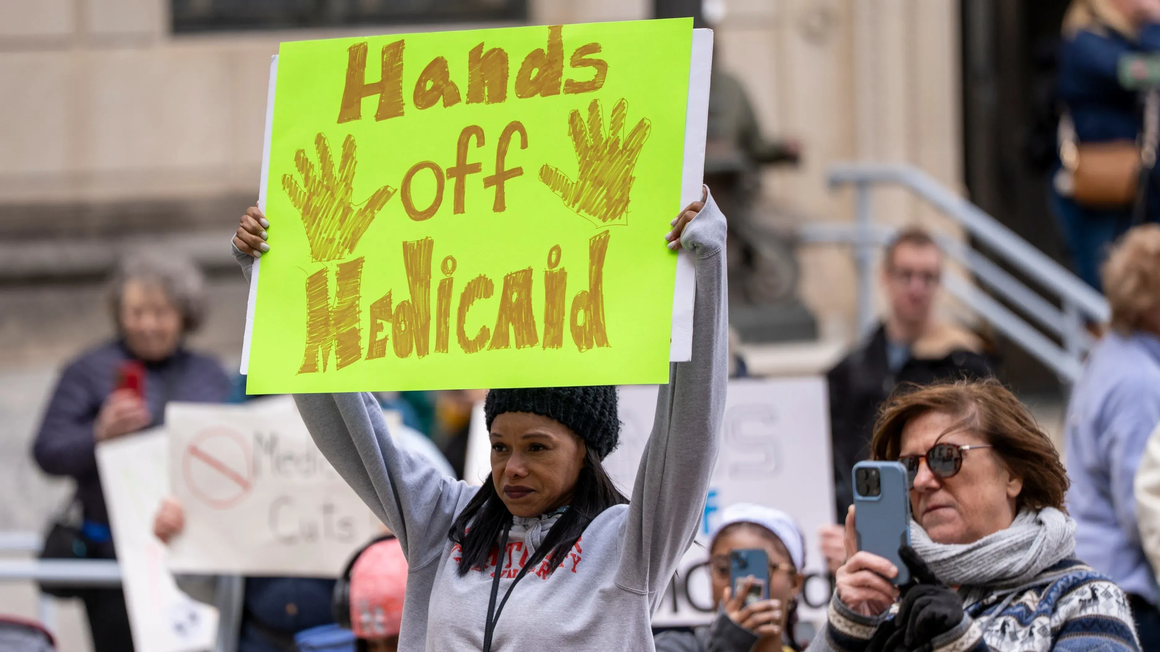 امرأة تحمل لافتة مكتوب عليها "أيدي بعيدة عن Medicaid"، بينما يتجمع المتظاهرون في احتجاج ضد تخفيضات برنامج Medicaid.