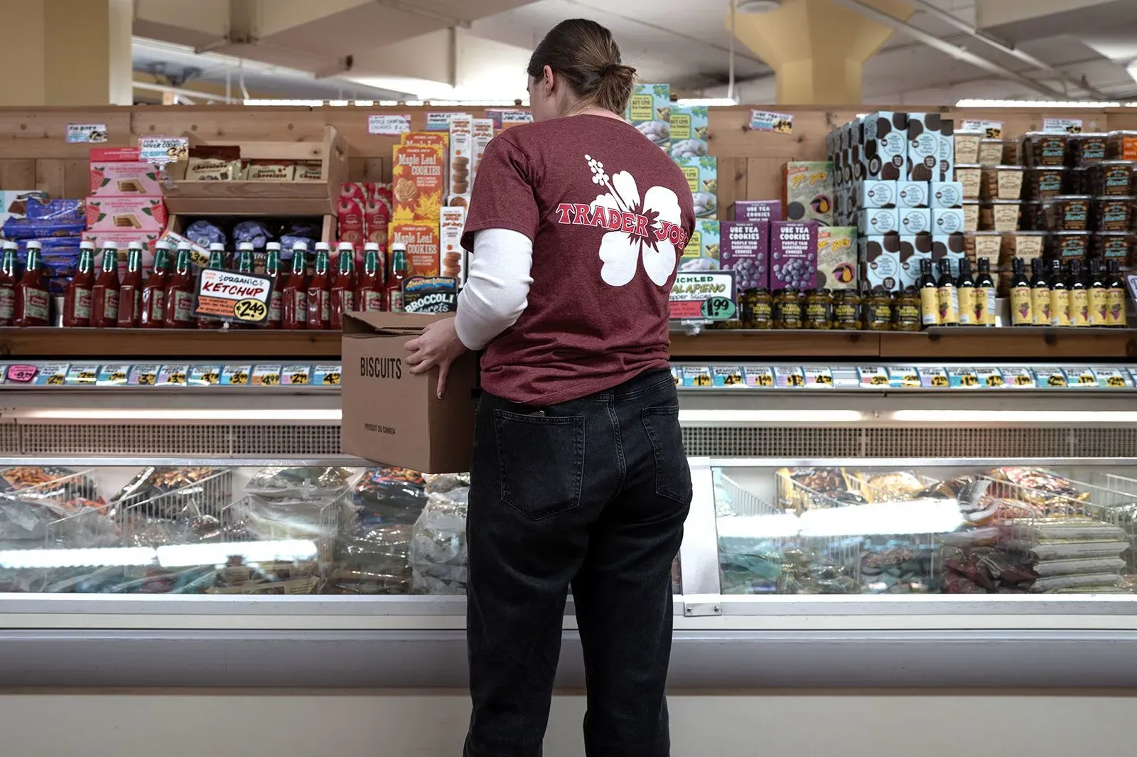 موظف في متجر "تريدر جو" يرتدي قميصًا يحمل شعار المتجر، يقوم بتحميل صناديق من البسكويت في قسم المجمدات، وسط مجموعة متنوعة من المنتجات الغذائية.