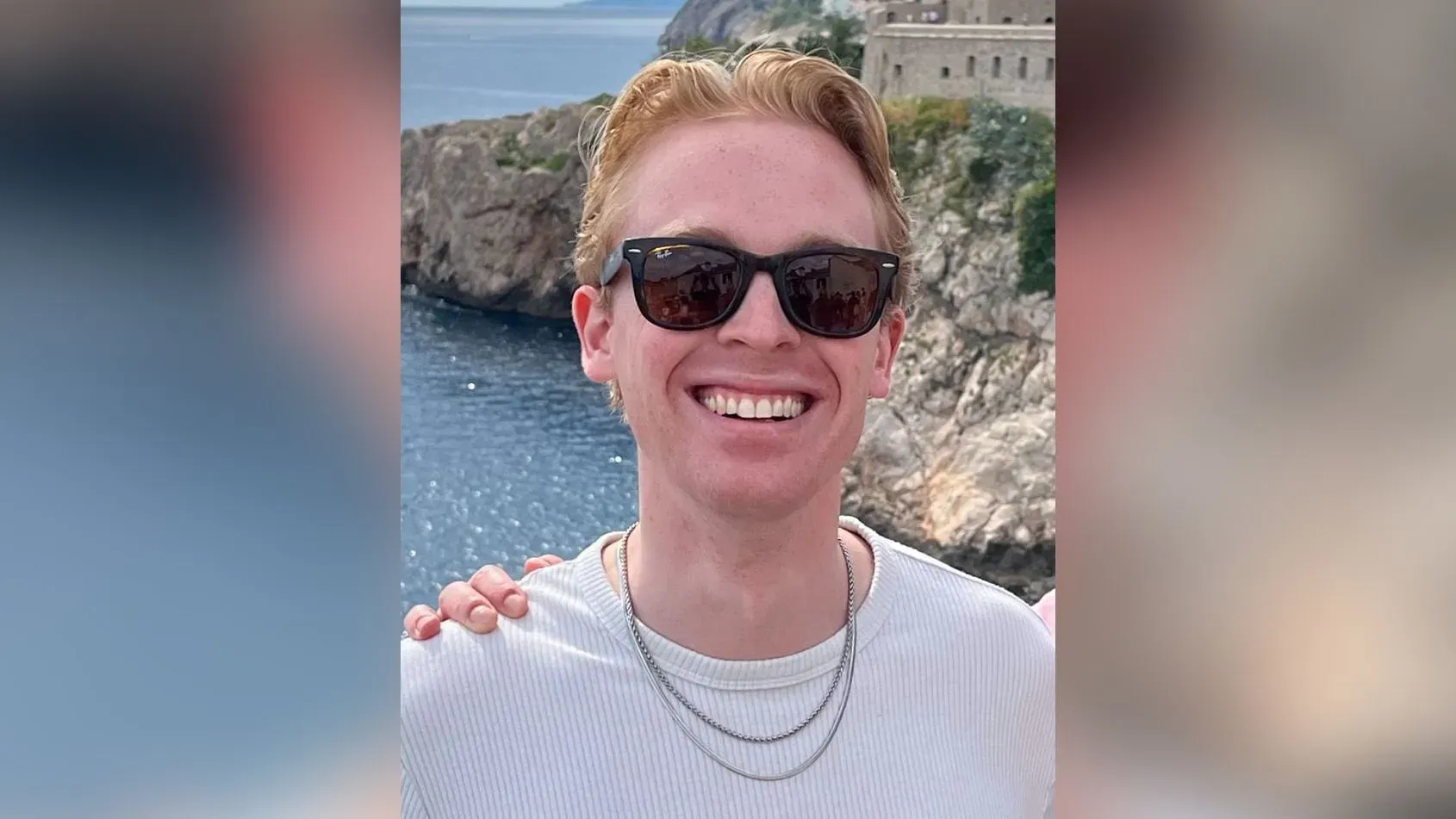 صورة لشاب مبتسم يرتدي نظارات شمسية، يظهر في خلفيته مناظر طبيعية جبلية، تعكس روح المغامرة والتنزه في الهواء الطلق.