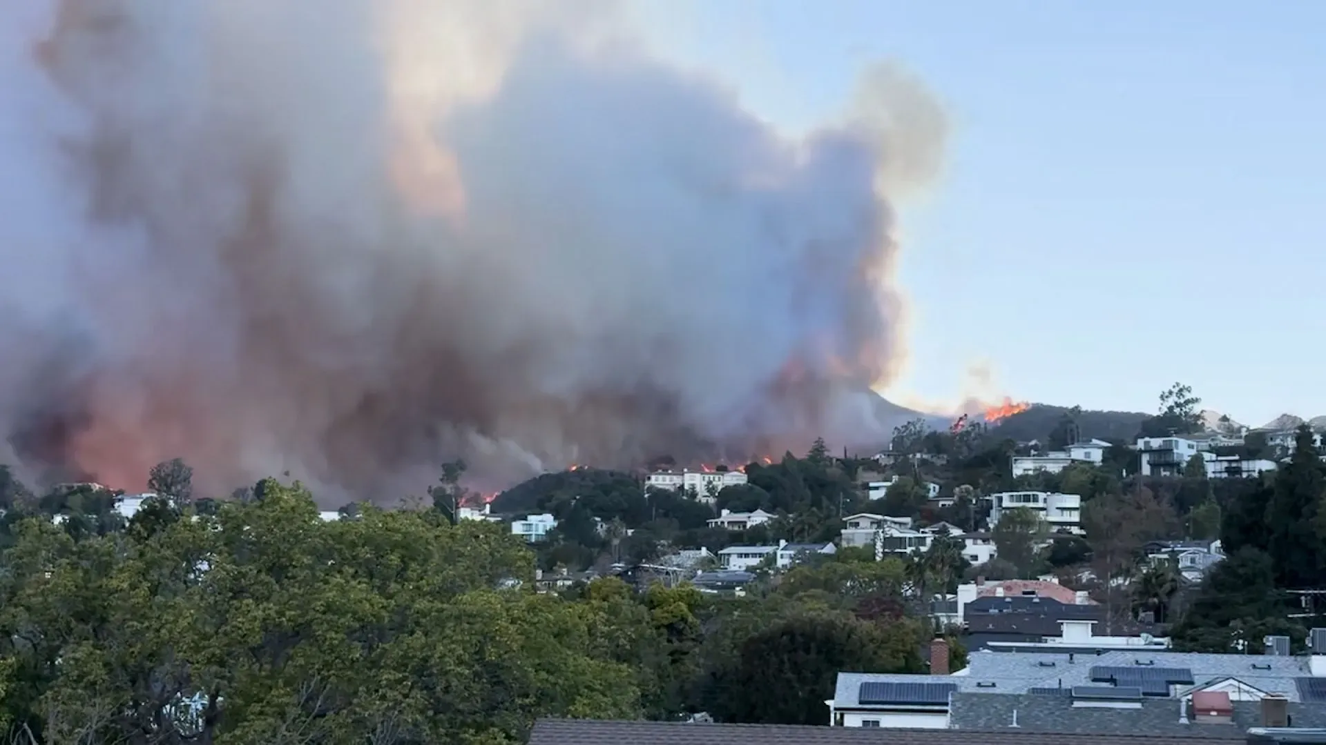 حرائق غابات تلتهم الجبال المحيطة بلوس أنجلوس، مع تصاعد الدخان فوق المنازل، مما يعكس تأثير الكارثة على المجتمع المحلي.