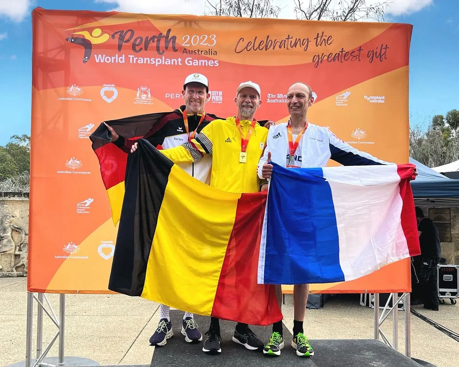 ثلاثة رياضيين يحملون أعلام بلدانهم (بلجيكا، ألمانيا، فرنسا) في دورة الألعاب العالمية لزراعة الأعضاء 2023 في بيرث، أستراليا، مع ميدالياتهم.