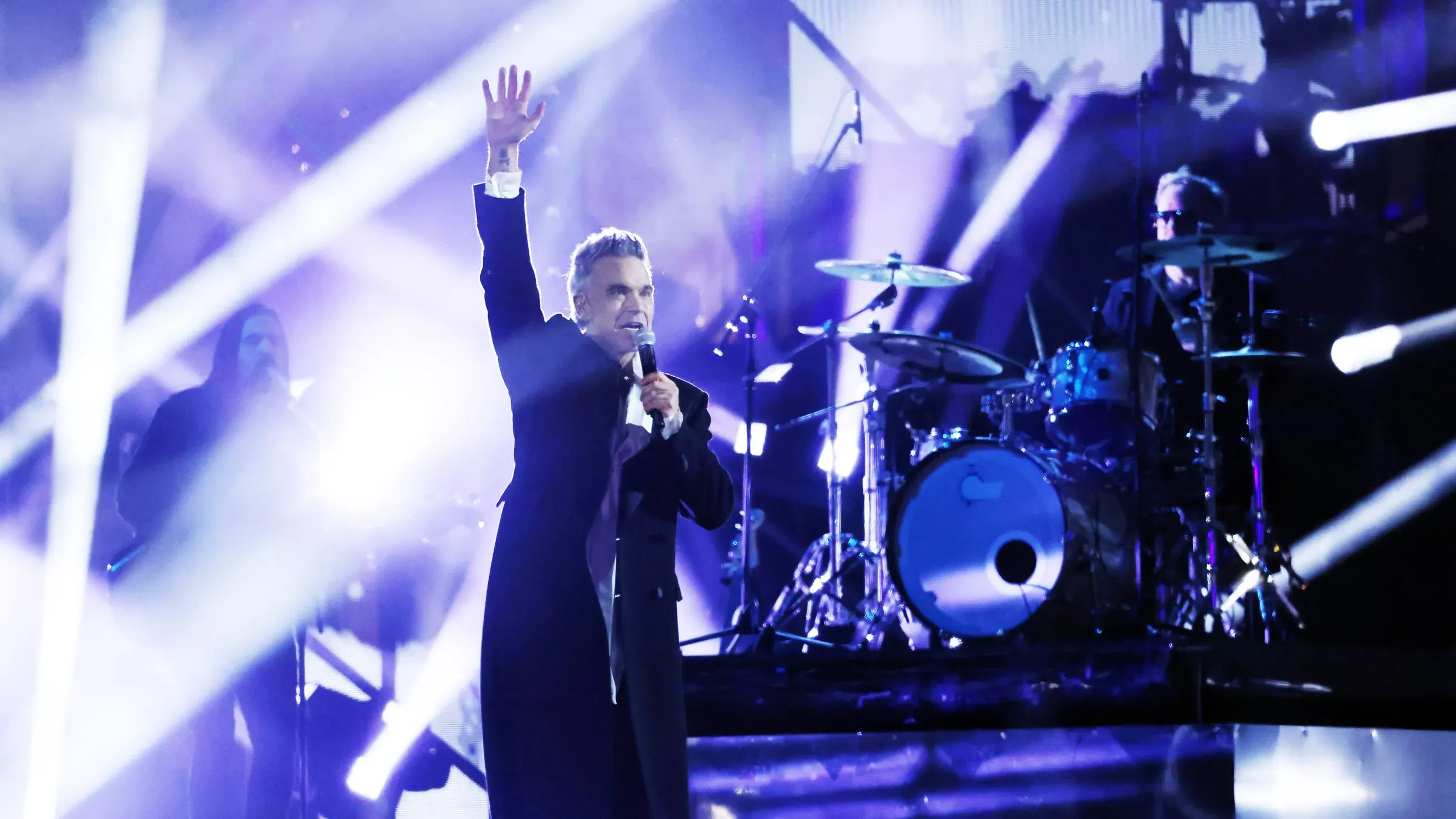 روبي ويليامز يؤدي على المسرح، مع إضاءة ساطعة وأعضاء فرقة خلفه، حيث يتحدث عن تحديات صحته النفسية ونقص فيتامين C.