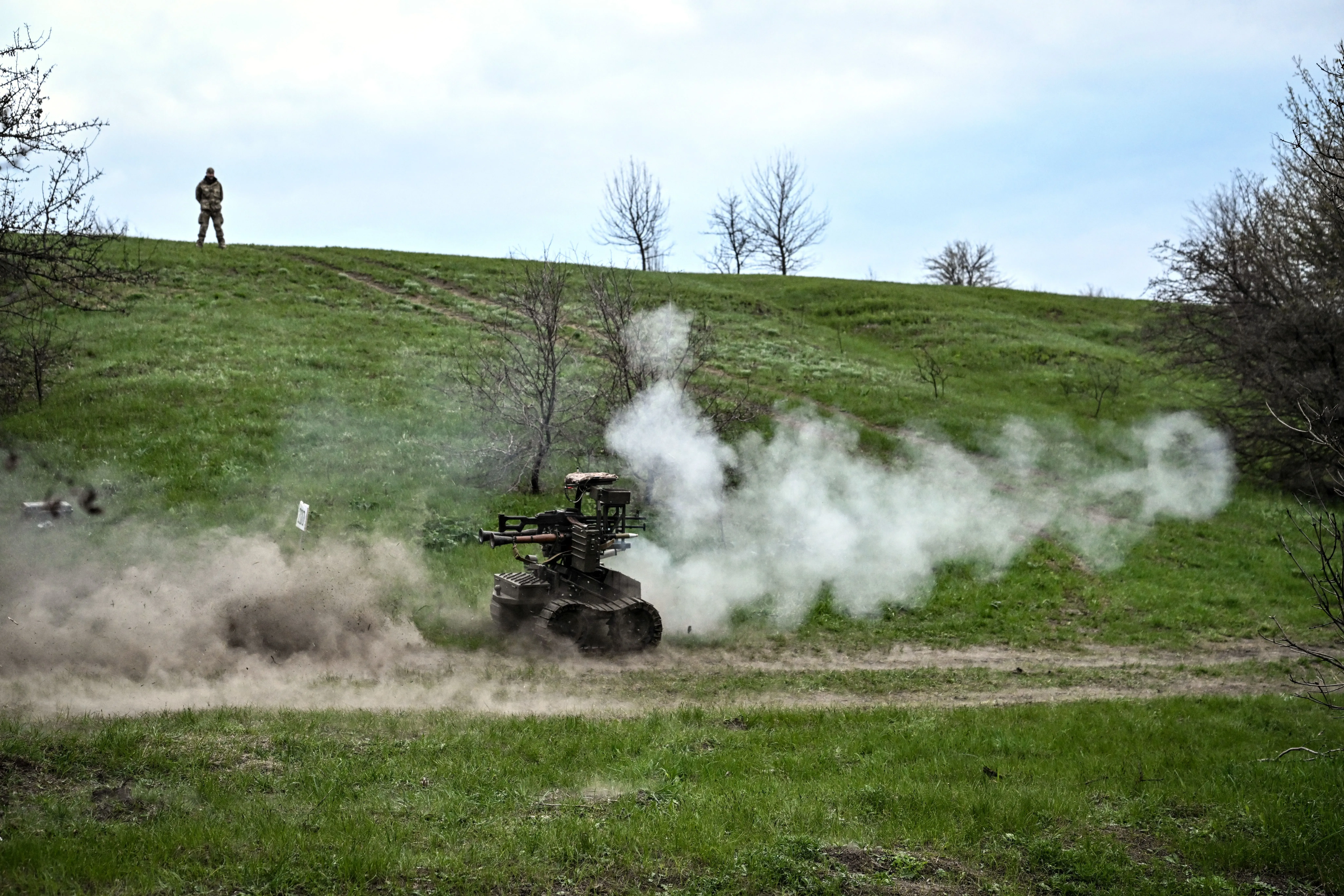 روبوت عسكري يطلق دخانًا أثناء تنفيذ مهمة في منطقة خضراء، مع جندي يقف في الخلفية، مما يعكس استخدام التكنولوجيا الحديثة في الحرب الأوكرانية.