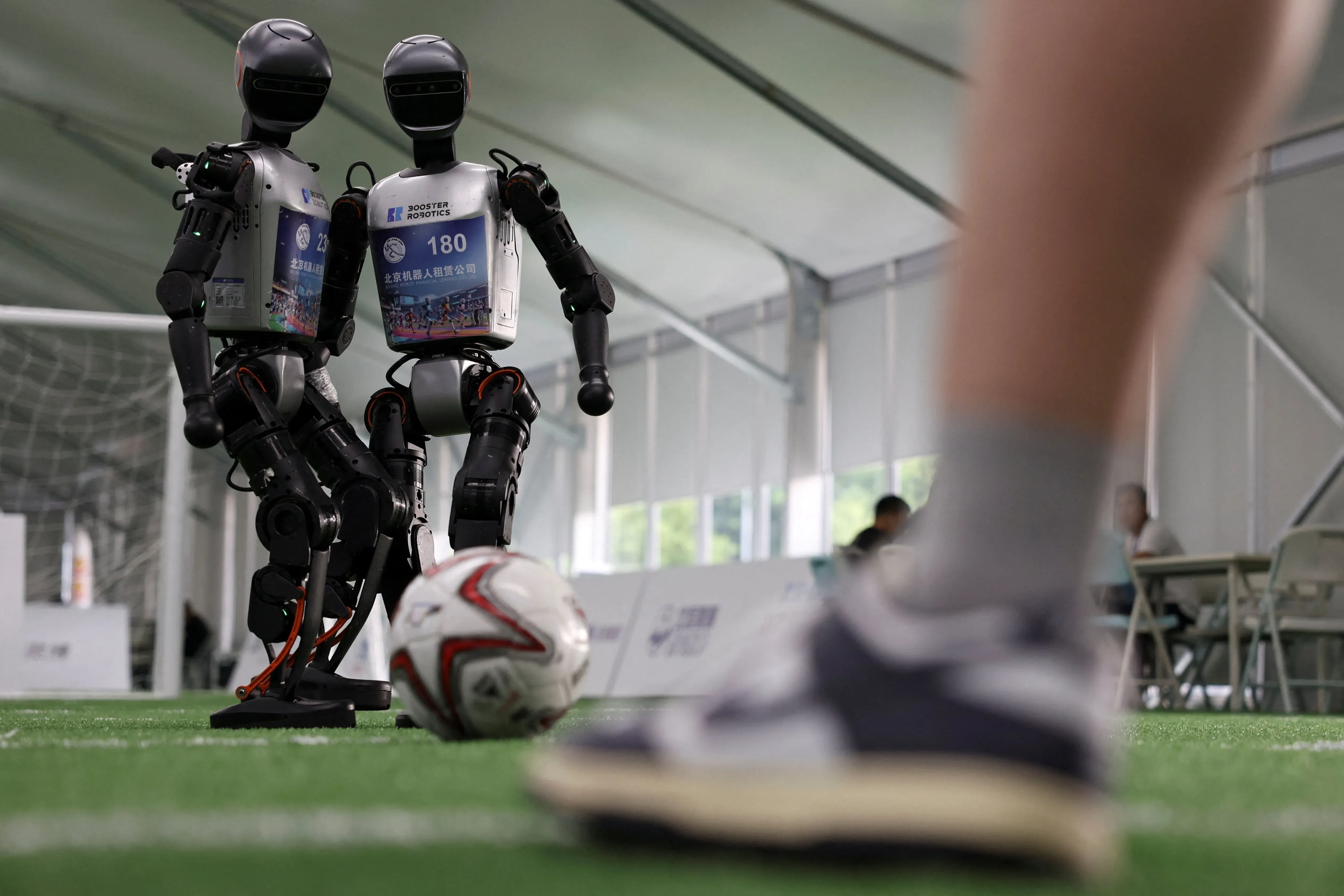 روبوتات تلعب كرة القدم في دورة الألعاب العالمية للروبوتات في الصين، مع كرة على الأرض، مما يعكس تقدم الذكاء الاصطناعي.