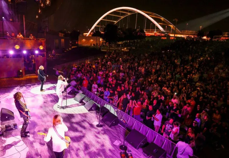 حفل موسيقي في الهواء الطلق يضم فرقة تؤدي على المسرح أمام حشد كبير، مع إضاءة ملونة وجسر في الخلفية.