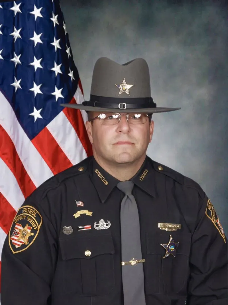 صورة لنائب مأمور شرطة أوهايو لاري هندرسون، يرتدي زي الشرطة الرسمي، مع العلم الأمريكي خلفه، في سياق قضية قتل.