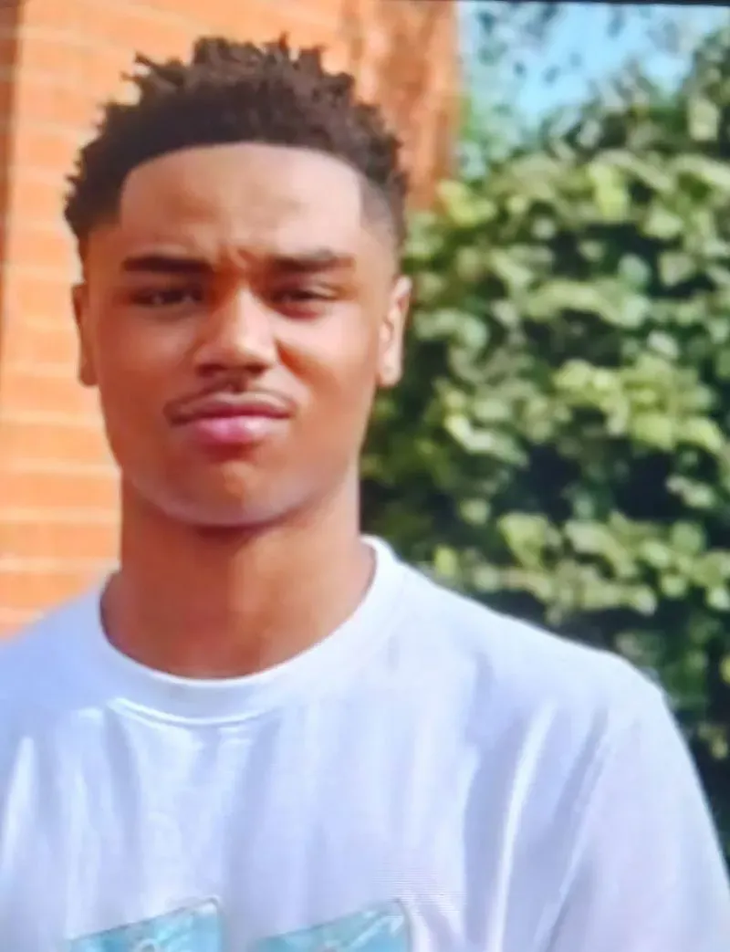 شاب مبتسم يرتدي قميصًا أبيض، يقف أمام خلفية خضراء، يمثل جزءًا من قضية تتعلق بمقتل ابنه برصاص الشرطة.