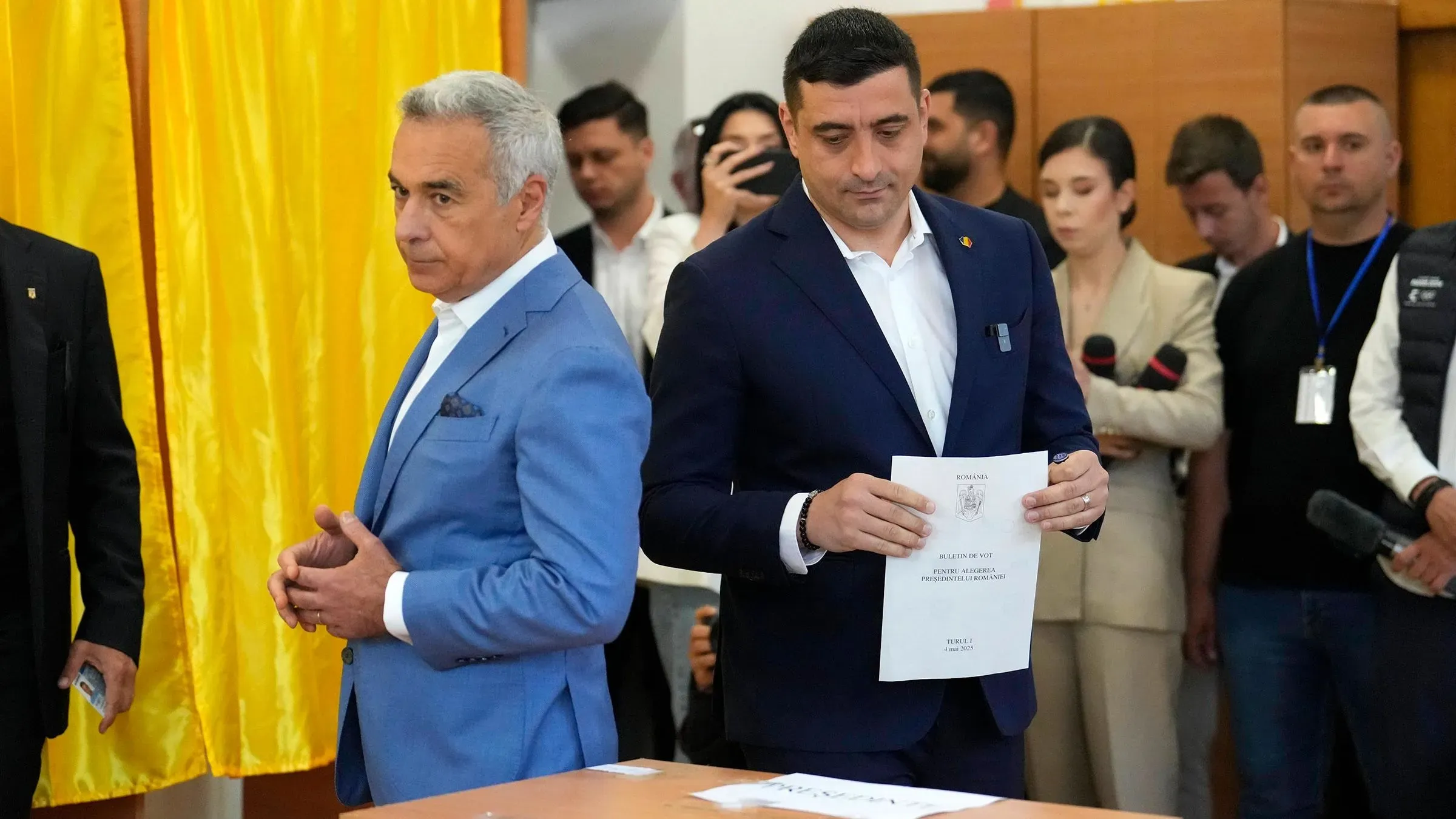 شخصان يقفان أمام ستارة اقتراع في مركز انتخابي، يحمل أحدهما ورقة اقتراع، بينما يتواجد آخرون في الخلفية.