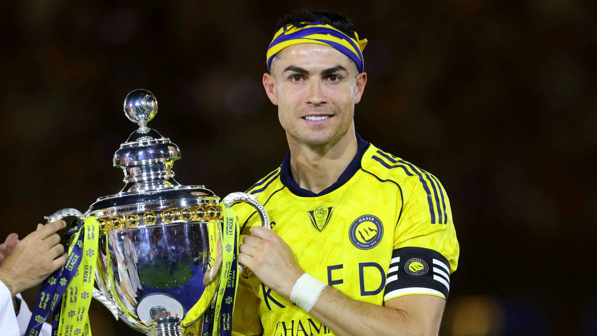 كريستيانو رونالدو قائد النصر السعودي يحمل كأس البطولة، مع التركيز على نهاية محتملة لمسيرته مع النادي وسط تحديات مالية وطموحات الفوز.