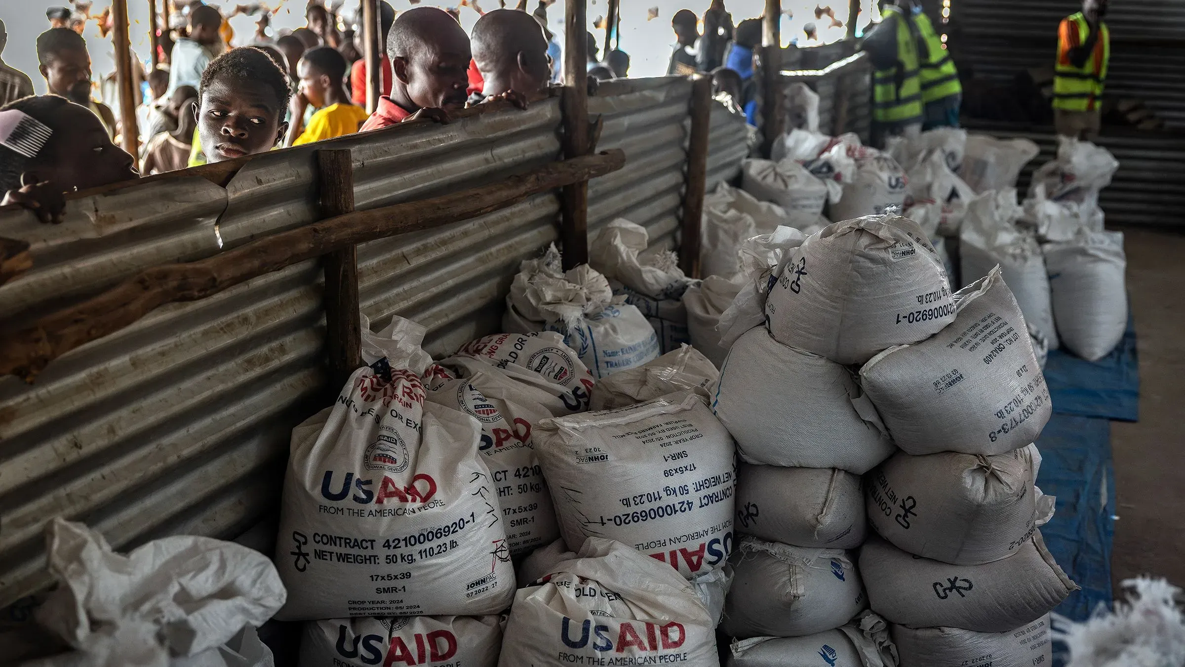 تظهر الصورة مجموعة من أكياس المساعدات التابعة للوكالة الأمريكية للتنمية الدولية (USAID) في مركز توزيع، مع وجود أشخاص في الخلفية ينتظرون الحصول على المساعدات.