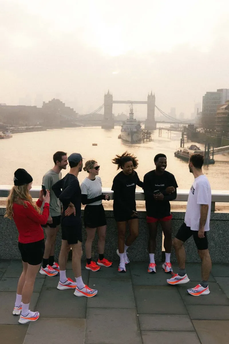 مجموعة من العدائين يرتدون ملابس رياضية عصرية، يتجمعون على ضفة نهر مع جسر في الخلفية، يعكس تزايد شعبية الجري كنشاط اجتماعي.