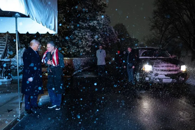 لقاء في الليل بين شخصين تحت مظلة، بينما تتساقط الثلوج. يظهر في الخلفية سيارة ومجموعة من الأشخاص، مما يعكس أجواء سياسية متوترة.