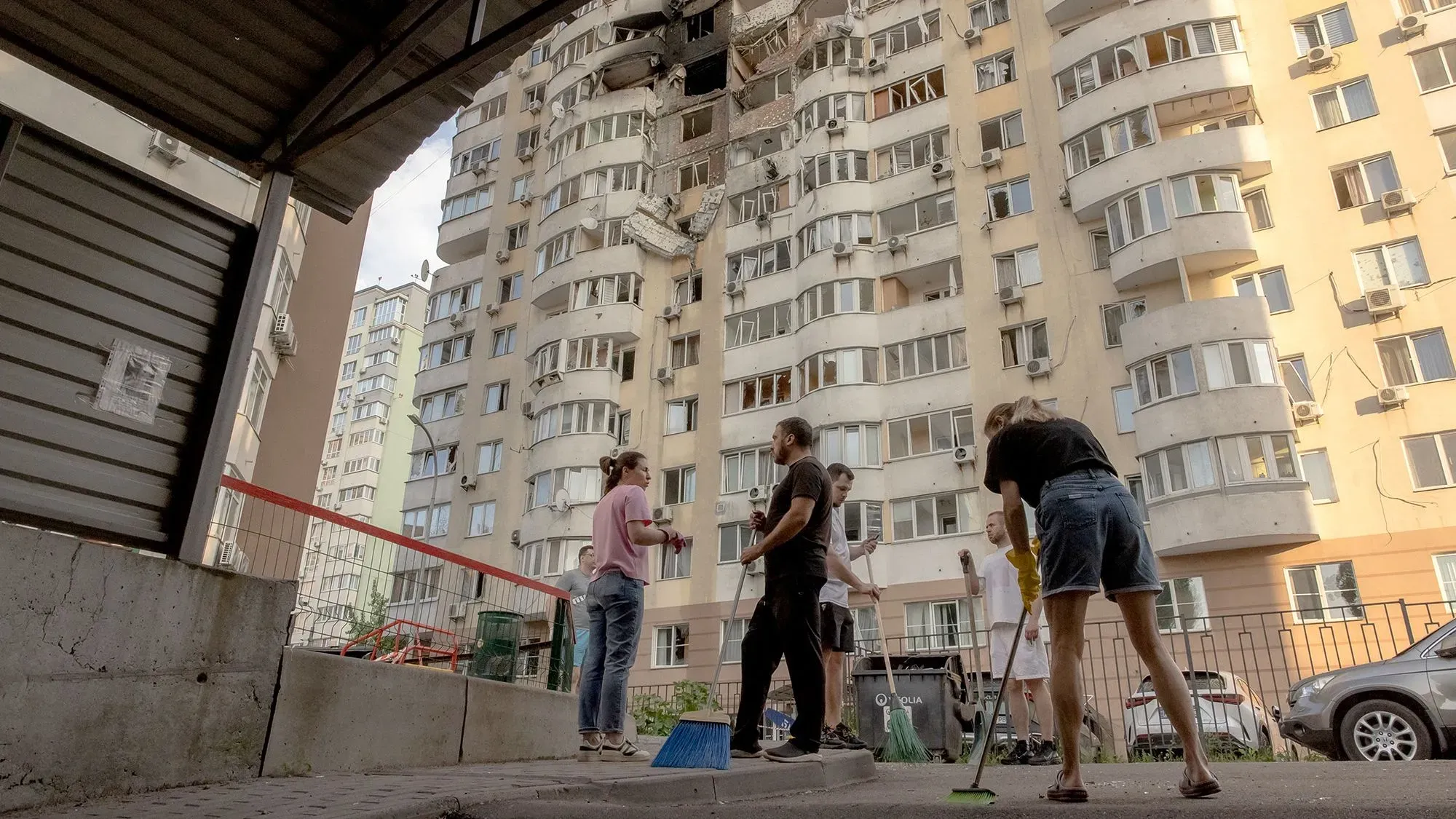 مجموعة من الأشخاص يقومون بتنظيف الفناء أمام مبنى سكني في كييف، حيث تظهر علامات دمار على الواجهة نتيجة الهجمات الجوية.