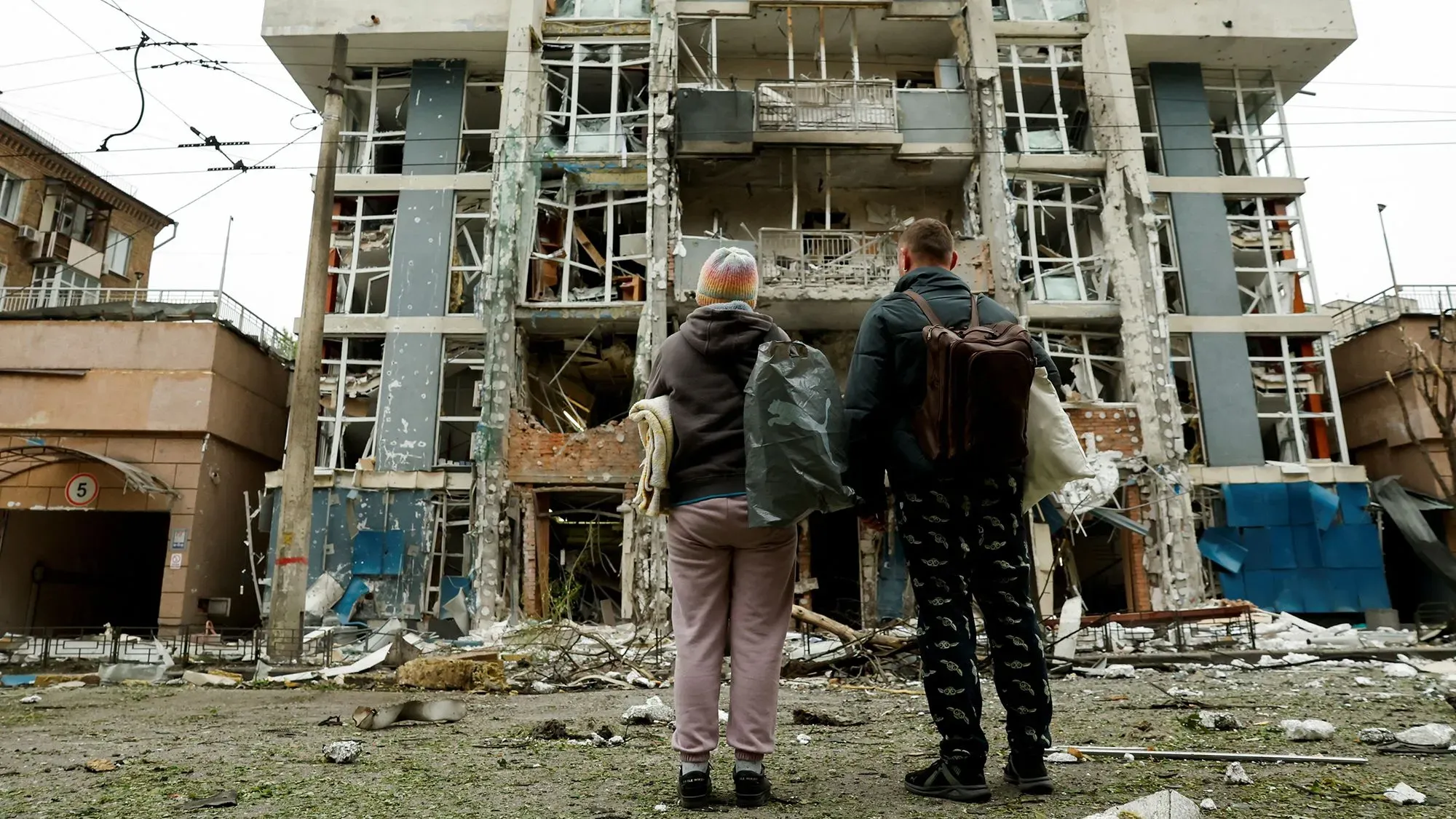 امرأتان تقفان أمام مبنى مدمر بفعل القصف، حيث تظهر آثار الدمار الشديد على الواجهة، مما يعكس تداعيات الحرب في أوكرانيا.