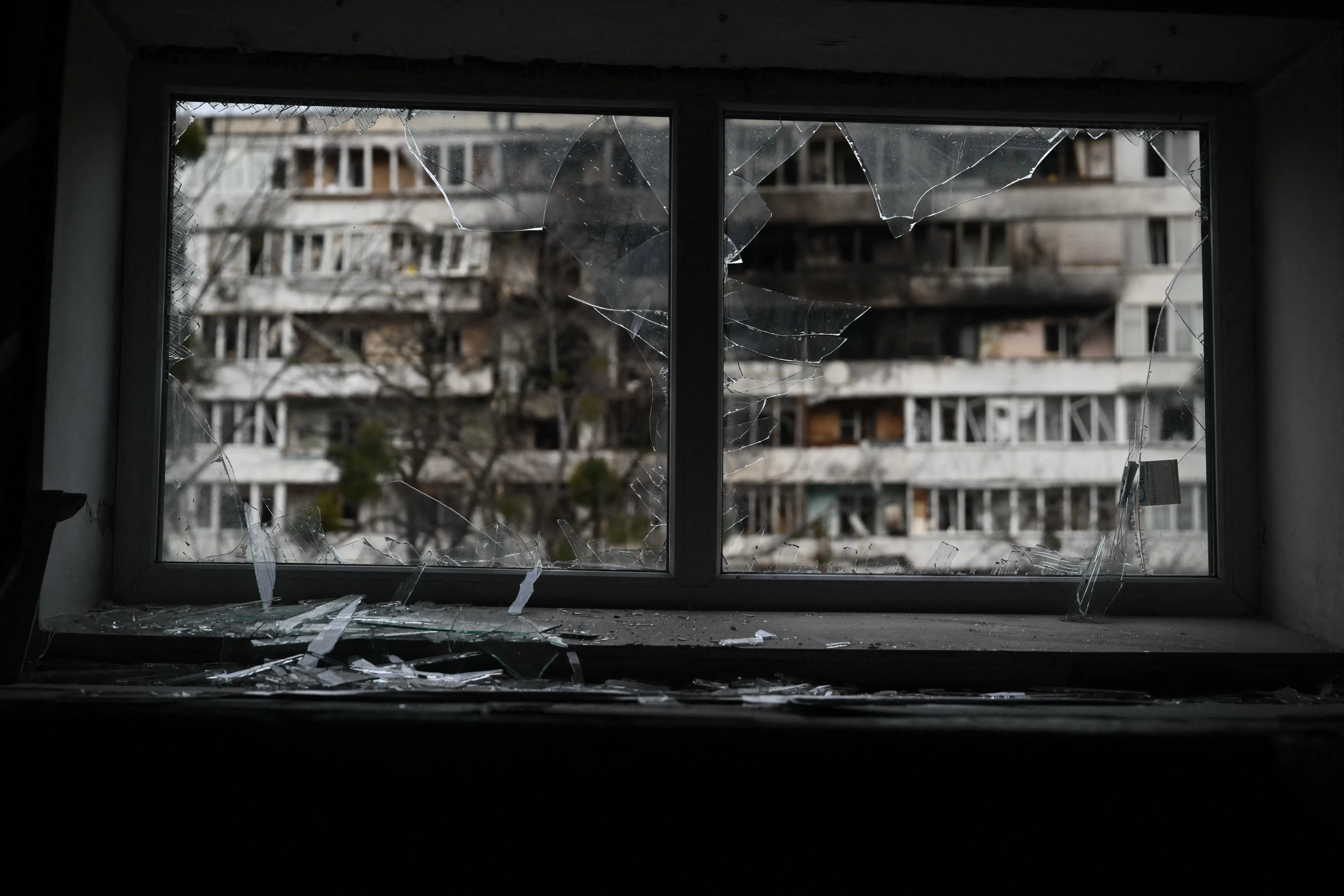 شباك مكسور يطل على مباني مدمرة في أوكرانيا، يعكس آثار الحرب والدمار الذي خلفته الضربات الجوية.