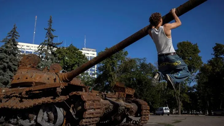 شاب يتدلى من مدفع دبابة قديمة في ساحة عامة، تعكس الصورة آثار الحرب والتوترات الجارية في أوكرانيا.