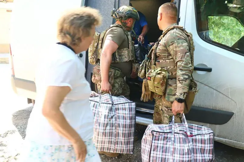 مدني يحمل حقائب كبيرة بينما يقوم جنود بتحميل الإمدادات في شاحنة، مما يعكس الوضع الإنساني الصعب في أوكرانيا خلال النزاع.