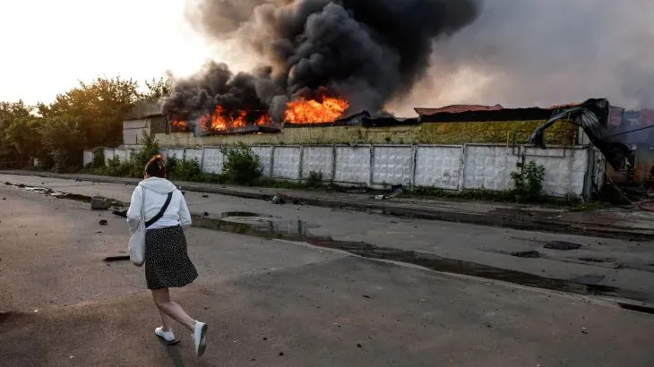 امرأة تسير نحو مبنى محترق، مع تصاعد الدخان الأسود والنيران، في مشهد يعكس آثار الهجمات الروسية على أوكرانيا.