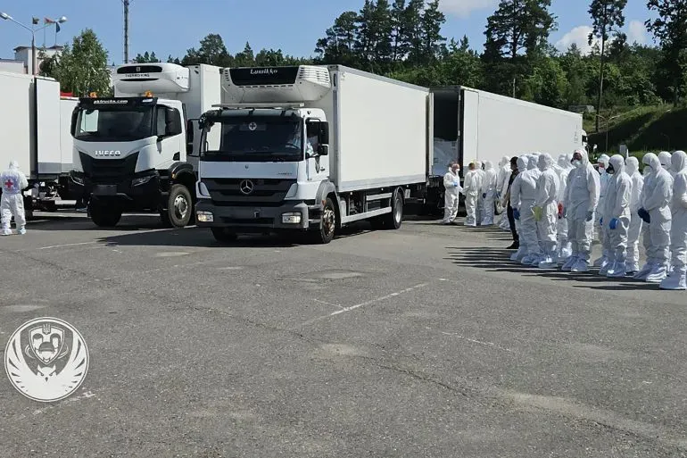 شاحنات نقل كبيرة متوقفة في ساحة، مع مجموعة من الأشخاص يرتدون بدلات واقية، وسط أشجار في الخلفية.