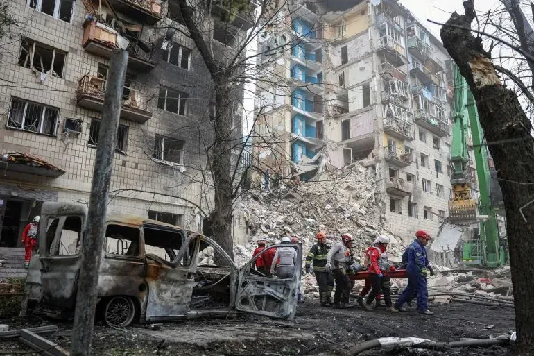 مشهد لفرق الإنقاذ تعمل على إجلاء الضحايا من موقع هجوم صاروخي في كييف، مع مباني مدمرة وسيارة محترقة في الخلفية.