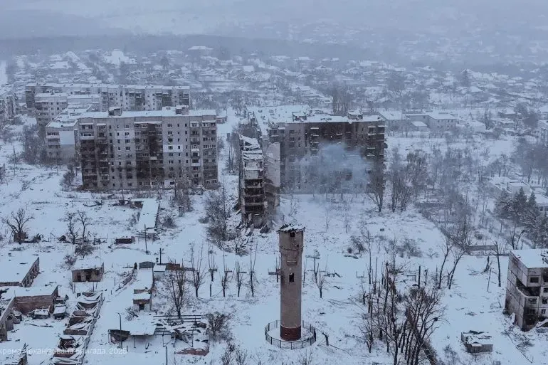 منظر لبلدة متضررة في أوكرانيا، تغطيها الثلوج، مع مباني مدمرة وأبراج مياه، تعكس آثار الصراع المستمر.