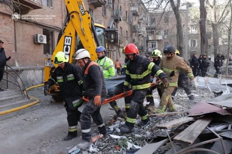 فرق الإنقاذ تعمل على نقل مصاب من موقع قصف روسي في أوكرانيا، مع وجود حطام في الخلفية وأفراد آخرين يتابعون الوضع.