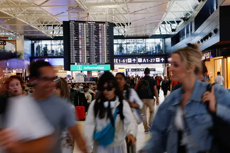 ازدحام في مطار دولي مع مسافرين يتجولون، وشاشة تعرض معلومات الرحلات والمغادرات. تعكس الصورة تأثير قيود السفر على السياح الروس.