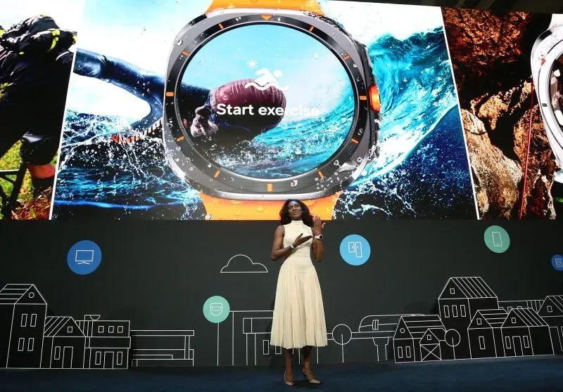 امرأة ترتدي فستانًا أبيض تتحدث أمام شاشة تعرض ساعة ذكية جديدة من سامسونج مع خيارات لممارسة الرياضة.