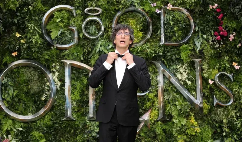 نيل غايمان، مؤلف \"Good Omens\"، يتحدث في حدث رسمي، وسط خلفية مزهرة تحمل اسم العمل، بعد مزاعم الاعتداء الجنسي ضده.