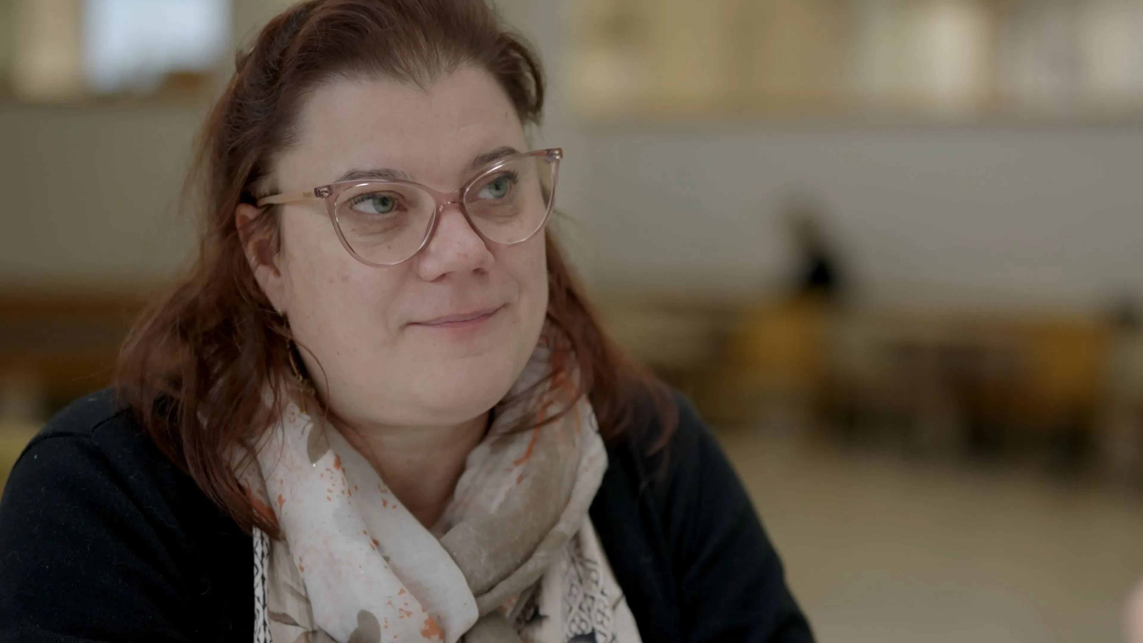 امرأة ترتدي نظارات وقميصًا أسود، تجلس في مكان عام، تعبر عن مشاعر القلق بشأن تأثير هجمات الذئاب على رعي الرنة في فنلندا.